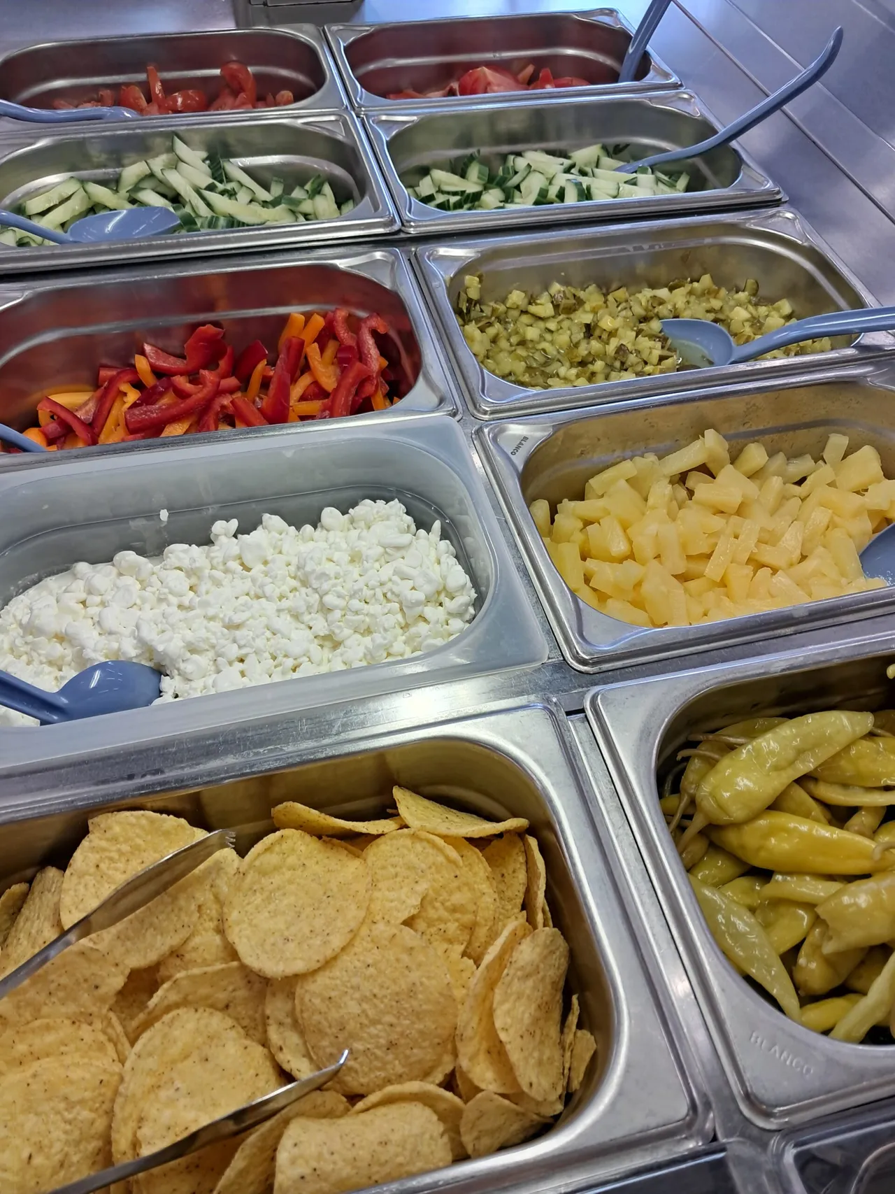 Close-up of a stainless-steel salad bar with trays of sliced tomatoes, cucumber, red and yellow bell peppers, diced pickles, pineapple chunks, cottage cheese, tortilla chips and pickled peppers.