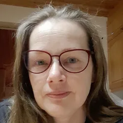 Close-up portrait of an adult woman with long brown hair and red-rimmed round glasses, neutral expression, soft lighting, standing in a home kitchen with wooden cabinets visible behind her.